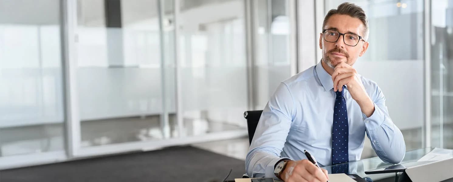 Mężczyzna w garniturze siedzi przy biurku w nowoczesnym biurze, patrzy zamyślony i trzyma długopis w dłoni.