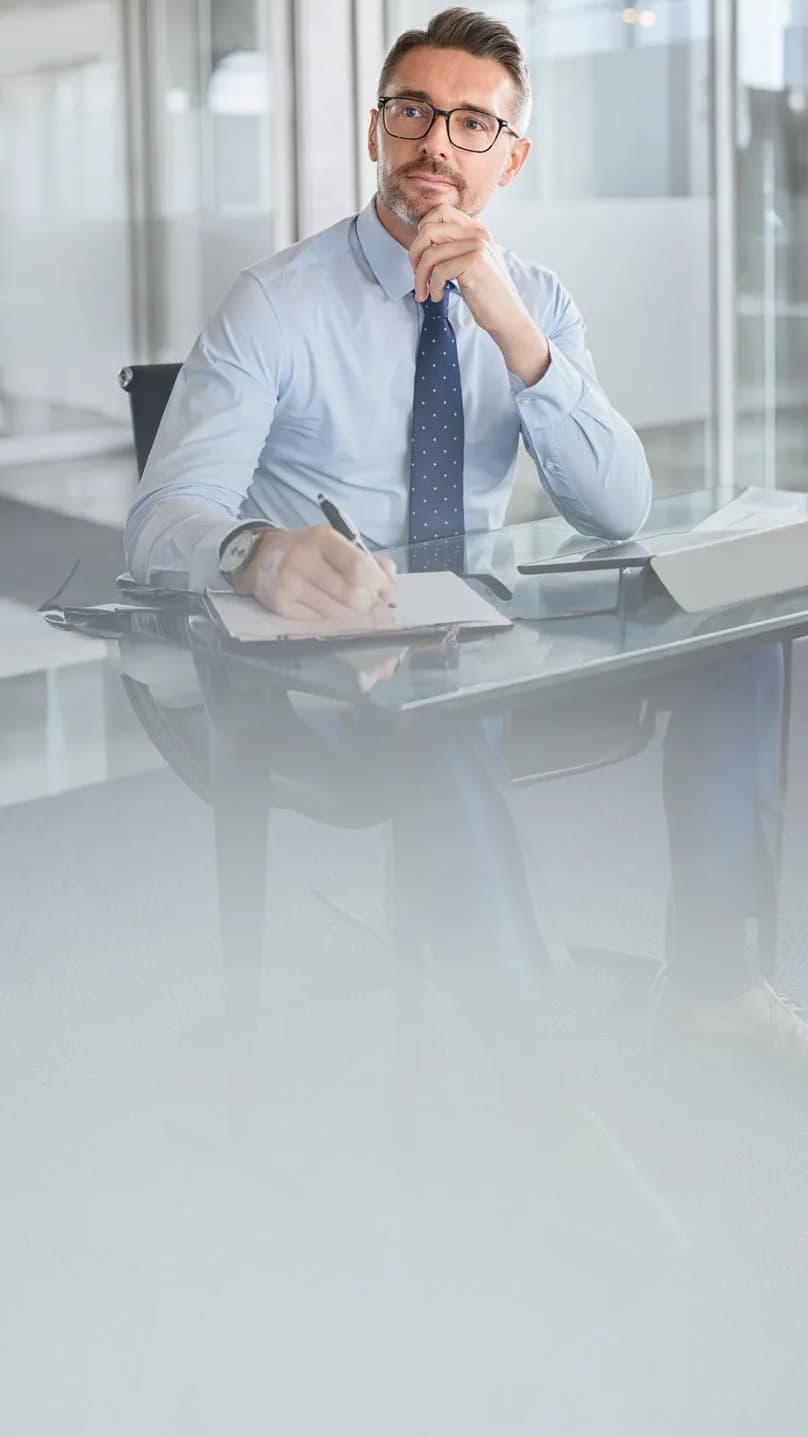 Mężczyzna w garniturze siedzi przy biurku w nowoczesnym biurze, patrzy zamyślony i trzyma długopis w dłoni.